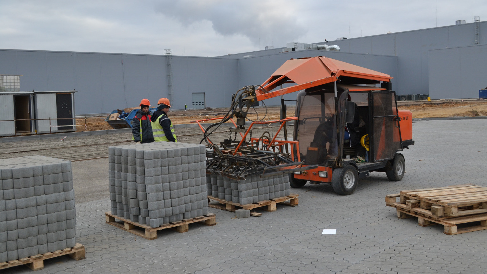 Two workers in safety gear observe a block-laying machine stacking concrete blocks on a construction site – Optimas.