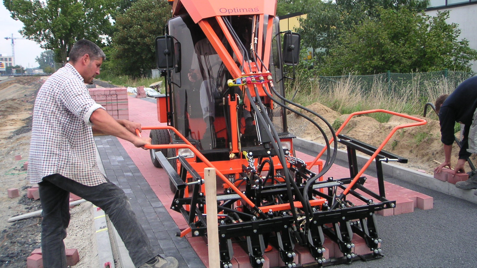 A worker operates a brick laying machine on a construction site, with bricks being placed on the ground – Optimas.