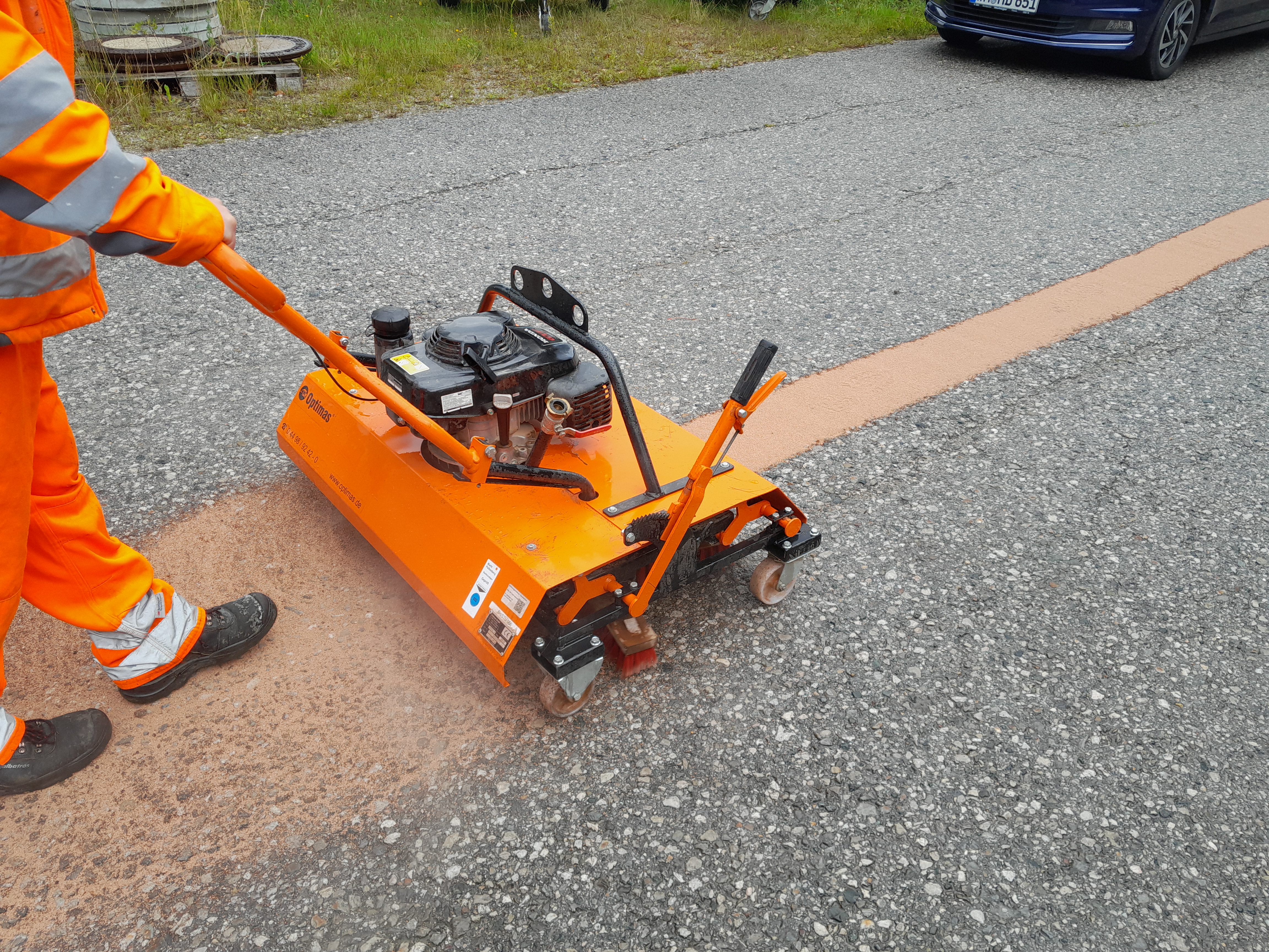 A worker in orange safety gear operates a line marking machine on a gravel road, creating a straight orange line – Optimas.
