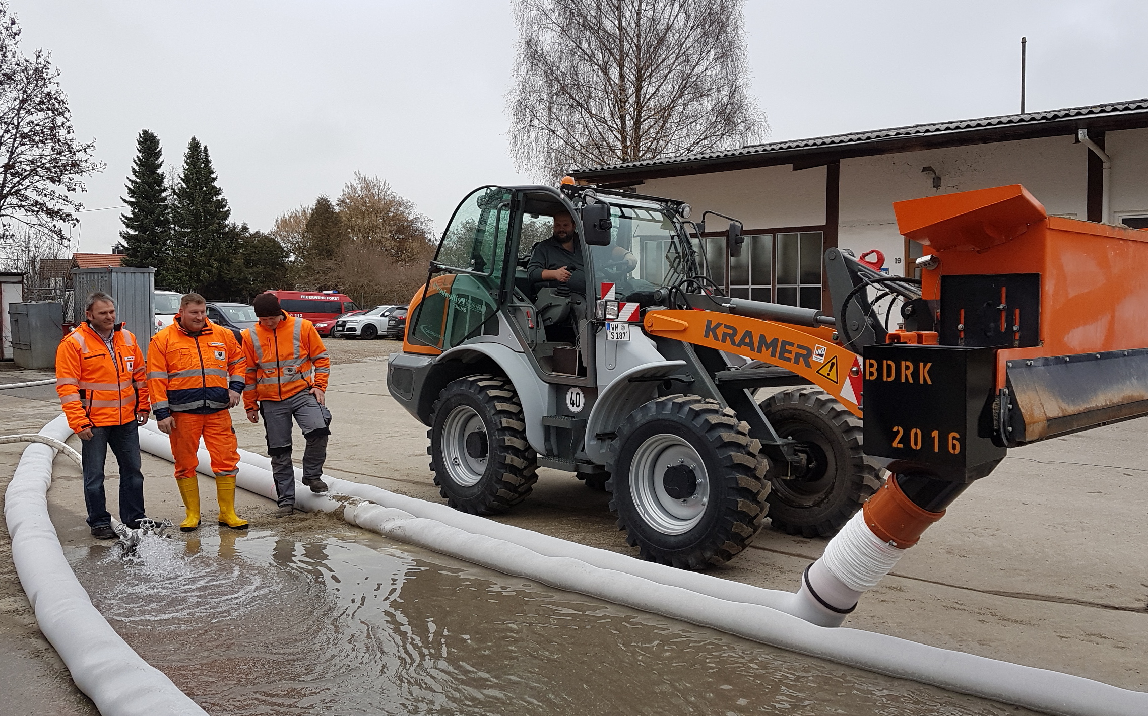 Ein Arbeiterteam testet ein Wasserableitungssystem mit einem großen orangefarbenen Kramer-Fahrzeug und weißen Rohrleitungen draußen in einer grauen und bewölkten Außenumgebung. – Optimas