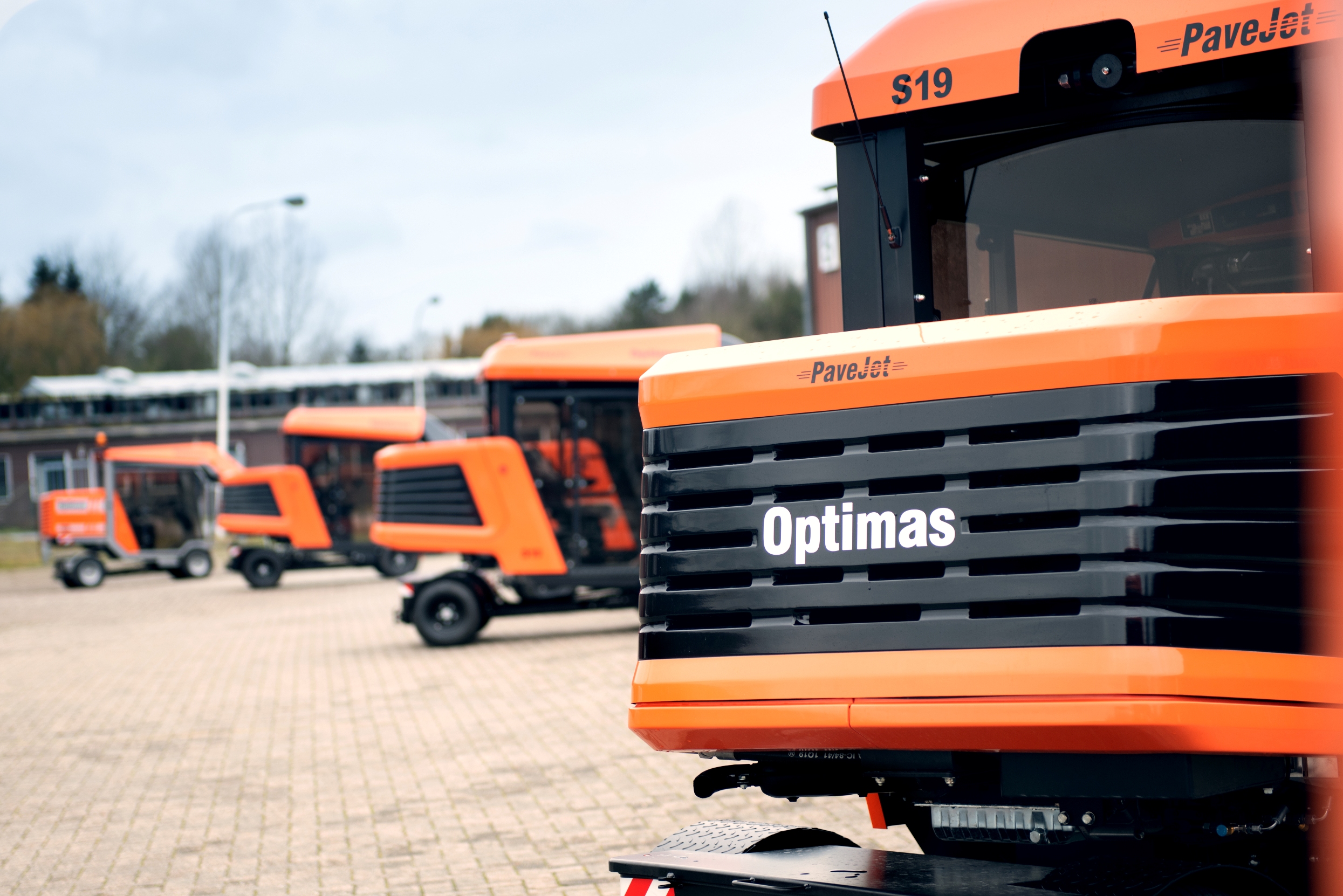 Eine Reihe orangefarbener Pflastermaschinen vom Typ PaveJet S19 stehen in einer Reihe im Freien und heben das „Optimas“-Logo auf der Vorderseite jeder Maschine hervor – Optimas.