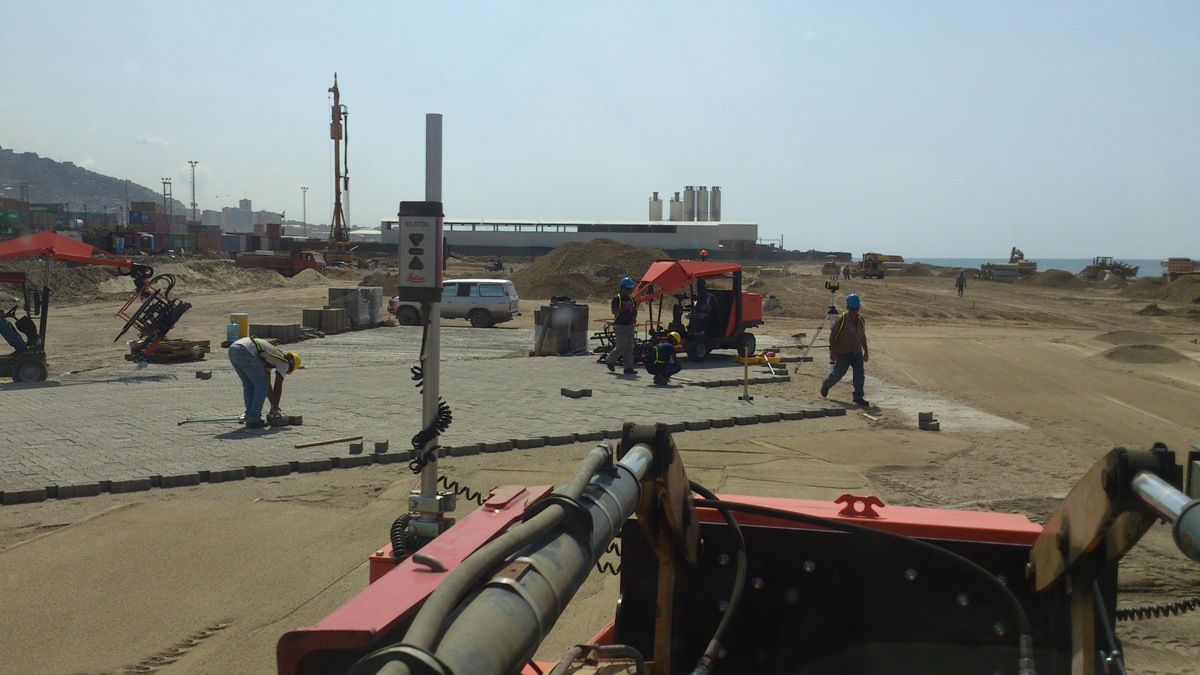 Construction workers laying pavement blocks at a beachside site with machinery in the background – Optimas.