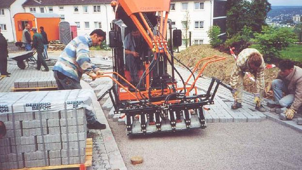 Workers using a paving machine to lay bricks on a construction site, with stacks of bricks nearby – Optimas.